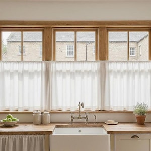 May include: A kitchen window with a wooden frame and three panes, each with a view of a stone building. White sheer curtains hang below the windows. A wooden countertop holds a white sink, jars, and a bowl of green apples.