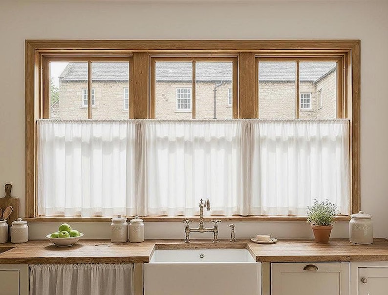 May include: A kitchen window with a wooden frame and three panes, each with a view of a stone building. White sheer curtains hang below the windows. A wooden countertop holds a white sink, jars, and a bowl of green apples.