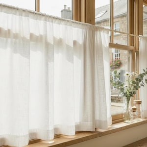 May include: Sheer white linen curtains hanging in front of a window. The curtains are semi-transparent, allowing sunlight to filter through. A wooden window frame and sill are visible, along with a view of a building and greenery outside.