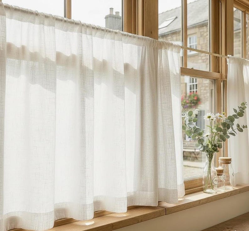 May include: Sheer white linen curtains hanging in front of a window. The curtains are semi-transparent, allowing sunlight to filter through. A wooden window frame and sill are visible, along with a view of a building and greenery outside.