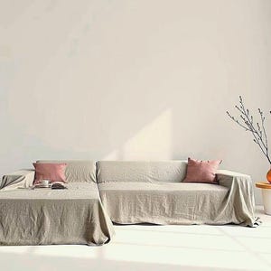 Könnte beinhalten: Beiges Ecksofa mit Leinenbezug und zwei rosa Kissen. Ein kleiner Beistelltisch mit einer Vase mit Zweigen. Der Raum hat ein minimalistisches Design mit natürlichem Licht.