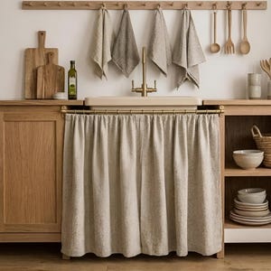 Puede incluir: Una cortina de tela beige para fregadero cuelga de una barra de latón debajo de un fregadero de cocina. El fregadero cuenta con un grifo de latón, rodeado de armarios de madera, paños de cocina colgantes, tablas de cortar y cuencos de cerámica.
