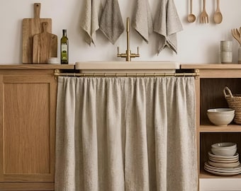 May include: A beige fabric sink skirt hangs from a brass rod beneath a kitchen sink. The sink features a brass faucet, and the surrounding area includes wooden cabinets, hanging kitchen towels, wooden cutting boards, and stacked ceramic bowls.