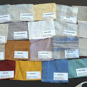 Puede incluir: Una colección de muestras de tela en varios colores, incluyendo gris, amarillo y azul. Cada muestra tiene una etiqueta con un número, una descripción y medidas en centímetros y gramos por metro cuadrado. Las telas están dispuestas sobre una superficie oscura.