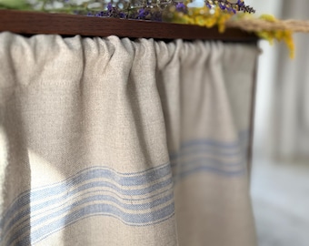 May include: A close-up of a linen fabric with blue horizontal stripes, draped over a wooden frame. The fabric is a light beige color. Purple and yellow flowers are placed on top of the wooden frame.
