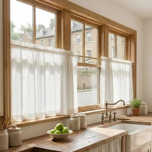 Könnte beinhalten: Eine Küche mit einer Holz-Arbeitsplatte, einem weißen Waschbecken und einem Fenster mit weißen Vorhängen. Die Fensterrahmen sind aus Holz. Eine Schale mit grünen Äpfeln steht auf der Theke, zusammen mit Gläsern und Utensilien.