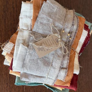 Puede incluir: Una pila de recortes de tela en varios tonos de marrón, naranja, rojo y verde, atados con cuerda. La tela es una mezcla de lino natural y tiene un aspecto rústico y hecho a mano.