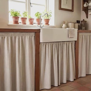 May include: A country-style kitchen featuring a white ceramic sink and a striped curtain. The sink is set into a wooden cabinet. Potted plants are on the windowsill above the sink. The curtains are beige with vertical stripes.
