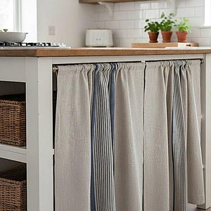 Peut inclure: Un ensemble de rideaux pour îlot de cuisine en aspect lin beige avec des rayures verticales bleu marine. Les panneaux de tissu sont froncés sur une tringle en métal, dissimulant l'espace de rangement d'un îlot blanc à plateau en bois.