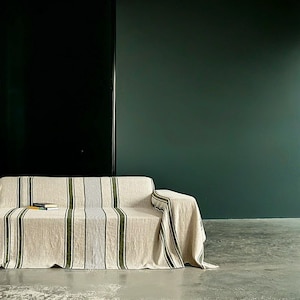 May include: A beige sofa with a striped cover featuring vertical green and black lines. The sofa is in front of a dark green wall and a black panel. Two books are on the sofa.
