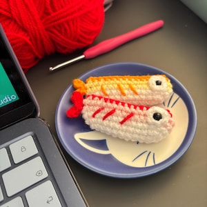 May include: Two small, crocheted fish-shaped ornaments resting on a round blue plate with a white cat face design. One fish is orange and white, while the other is red and white. Both feature black bead eyes and small, textured fins.