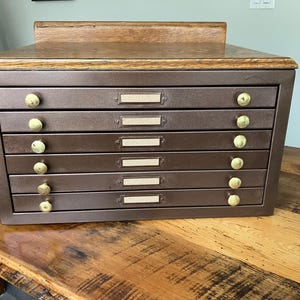 May include: A brown metal flat file cabinet with six drawers, each featuring a brass-colored knob and a rectangular label holder. The cabinet is topped with a finished wooden surface that has a raised back edge, providing a sturdy storage unit.