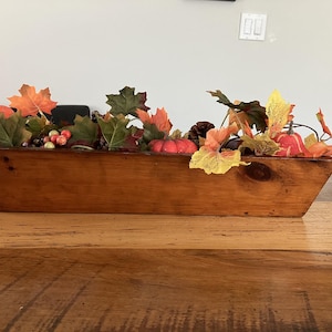 May include: A long, rectangular wooden dough bowl filled with artificial autumn foliage, including green and orange maple leaves, small red pumpkins, and clusters of berries. The bowl has a smooth, medium-brown wood grain finish.