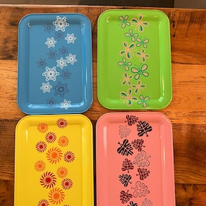 May include: Four rectangular serving trays in blue, green, yellow, and pink. Each tray features a different decorative design: snowflakes, butterflies, sunbursts, and leaves. The trays are arranged on a wooden surface.