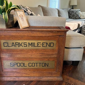 May include: A rectangular wooden storage cabinet with a natural brown wood grain finish. The front features two horizontal panels with the black text CLARK'S MILE-END and SPOOL COTTON printed on a muted gold background. The cabinet has a flat top surface.