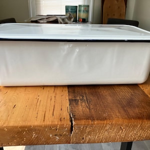 May include: A rectangular white enamel storage container with a matching lid and a thin black rim along the top edge. The container has smooth, slightly rounded corners and a glossy finish, presenting a clean and functional design for storage.