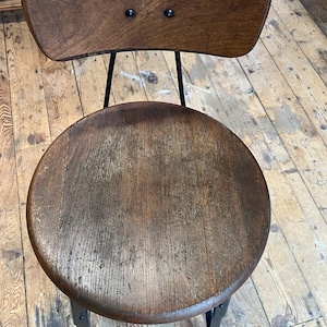 May include: A vintage wooden chair with a round seat and a small backrest. The seat and back are made of distressed wood, and the frame is black metal. The chair has a rustic, industrial aesthetic.