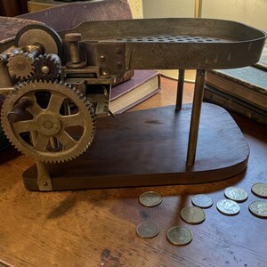 May include: A vintage metal coin sorting machine featuring a large gear, a perforated tray, and a wooden base. The mechanical device is constructed with exposed gears and a metal chute. Several metallic coins are scattered on the surface nearby.
