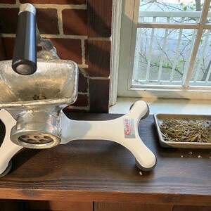 May include: A vintage, white and silver meat grinder with a black handle. The grinder has a funnel-shaped top and a circular grinding plate. The base is white with the brand name "Choprite" on it. The grinder is on a wooden surface.