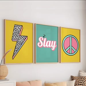 Op de afbeelding: Drie ingelijste wandkunstwerken. De eerste heeft een gele achtergrond met een bliksemschicht met zwart-witte luipaardprint. De tweede heeft het woord "Slay" in roze en wit op een turquoise achtergrond. De derde heeft een vredesteken op een gele achtergrond.