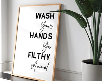 May include: A framed white print with the text "WASH YOUR HANDS YA FILTHY ANIMAL" in black script and block letters. The frame is light brown. A large green plant in a white pot is to the right of the print.