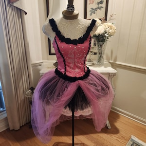 May include: A pink and black bodice dress featuring a sweetheart neckline with black ruffled trim, a central panel adorned with small silver studs, and a layered tulle skirt with a shorter black inner layer and a longer pink outer layer.