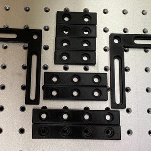May include: Black metal L-shaped brackets and rectangular connectors arranged on a silver surface with evenly spaced holes. A ruler with centimeter markings is visible along the right edge. The items appear to be for construction or assembly.