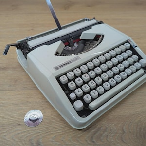 Op de afbeelding: Een lichtgrijze Hermes 3000 schrijfmachine. De schrijfmachine heeft een volledig toetsenbord met witte toetsen en zwarte letters. Het Hermes-logo is zichtbaar aan de voorkant. Er ligt een klein, rond zilveren embleem naast.