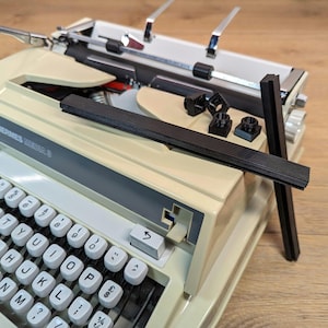 Pode incluir: Uma máquina de escrever vintage branca Hermes Meda 3 com uma capa de teclado de plástico preto e um conjunto de peças de plástico preto. A máquina de escrever tem um teclado preto e cinza com teclas etiquetadas com letras, números e símbolos.