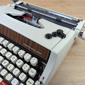 May include: A vintage Brother Deluxe 1310 typewriter with a cream-colored body and a wood-grain panel. The keyboard features white keys with black lettering. The text "AUTOMATIC REPEAT SPACER" is visible. Several black circular keys are on top.
