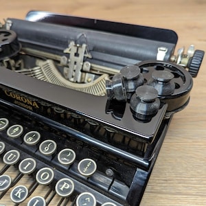 May include: A black Corona Junior typewriter with a black carriage return lever and a black metal frame. The typewriter has a black plastic roller and a black plastic knob. The typewriter has a black plastic keyboard with white keys and black lettering. The keys are labelled with letters, numbers, and symbols.