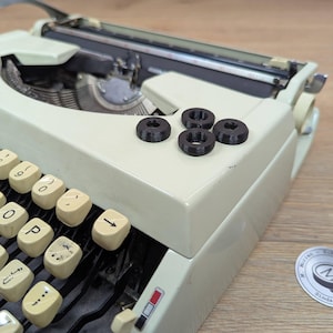 May include: A vintage off-white typewriter with cream-colored keys. Four small black circular objects are on the typewriter's surface. The typewriter is on a wooden surface. A sticker with text is in the lower right corner.