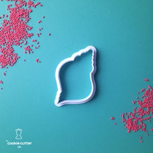 May include: A white plastic seashell-shaped cookie cutter on a teal surface, with scattered pink sprinkles. The cookie cutter has a scalloped edge. The logo for "cookie cutter lab" is in the lower left corner.