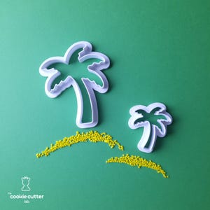 Könnte beinhalten: Zwei weiße Ausstecher in Form von Palmen auf grünem Hintergrund, mit gelben Streuseln, die wie Sand angeordnet sind. Die Ausstecher sind in verschiedenen Größen. Der Text "the cookie cutter lab." befindet sich unten links.