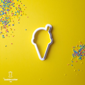 Puede incluir: Un cortador de galletas de plástico blanco con forma de cono de helado sobre un fondo amarillo. Hay chispas de colores esparcidas alrededor del cortador. Las palabras "cookie cutter lab" están impresas en la parte inferior.