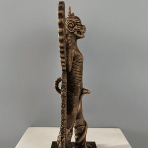 May include: A bronze statue of a mythical creature with a serpent wrapped around its leg. The creature has a human-like body with a feline head and wings. The statue is standing on a square base.
