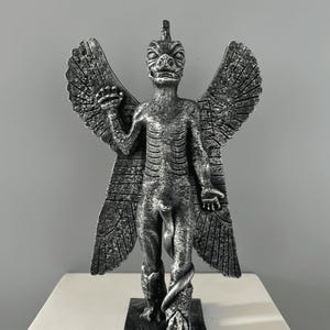 May include: A silver-colored statue of a winged demon with a snake wrapped around its legs. The demon has a reptilian head with a pointed crown and a human-like body. The statue is standing on a square base.