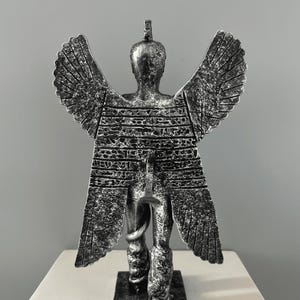 May include: A silver-colored statue of a winged figure with a snake wrapped around its leg. The figure is standing on a square base.