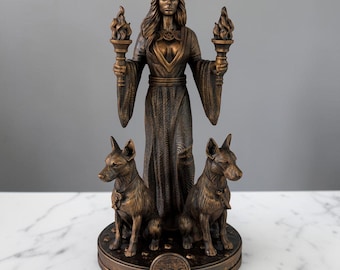 May include: A bronze-toned statue of a standing figure wearing a long, flowing robe, holding a flaming torch in each hand. Two dogs sit at the figure's feet on a circular base featuring a triple moon symbol. The figure wears a pentacle pendant.