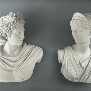 Può includere: Due busti in gesso bianco di figure classiche. Il busto a sinistra raffigura un uomo con capelli ricci e un drappeggio. Il busto a destra mostra una donna con un copricapo e abiti drappeggiati. Entrambi su sfondo grigio.