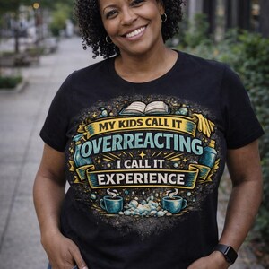 Pode incluir: Camiseta preta com o texto "MY KIDS CALL IT OVERREACTING I CALL IT EXPERIENCE" em um design decorativo. A camiseta apresenta um livro, xícaras de café e outros elementos gráficos. A pessoa está usando jeans azul.
