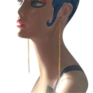 May include: Long, delicate gold chain earrings. The earrings are shown on a mannequin with blue hair and painted facial features. The chains hang down, creating a simple yet elegant look. The earrings are a classic accessory.