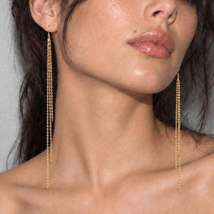 May include: A pair of long gold chain earrings featuring multiple delicate, thin, dangling metallic strands that hang vertically from the earlobe. The earrings consist of several fine, interconnected links that create a fluid, elongated appearance.