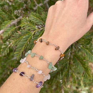 May include: A set of four bracelets with different colored stones, including amethyst, rose quartz, green aventurine, and tiger's eye. The bracelets are made with silver wire and feature small, irregular shaped stones.