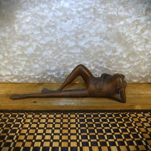 May include: Bronze frog sculpture in a relaxed pose, resting on its elbows with one leg bent. The frog has detailed features and is set against a textured background. The sculpture is a decorative item.