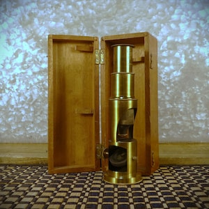 May include: A vintage brass microscope with a wooden case. The microscope is composed of several cylindrical sections and sits upright in the open wooden box. The box is made of light-colored wood and has brass hinges.