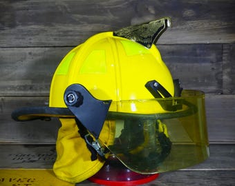May include: A bright yellow firefighter's helmet with a clear face shield and a gold-colored eagle emblem on top. The helmet has black hardware and a yellow neck protector. The helmet is sitting on a red stand.