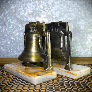 May include: Two bronze-colored Liberty Bell replicas. Each bell is mounted on a rectangular, marble-like base with brown and white veining. The bells have a detailed design and are set against a textured, metallic background.