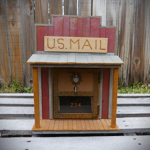 May include: A rustic wooden mailbox designed to resemble an old-fashioned building. The mailbox features a red and brown exterior with the words "U.S. MAIL" carved into a wooden sign. The door has a window with the number "234".