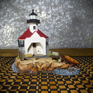 May include: A detailed miniature lighthouse model with a red roof, white walls, and a black lantern room. The model sits on a rocky base with a small boat and other nautical elements. The base has a small body of water.